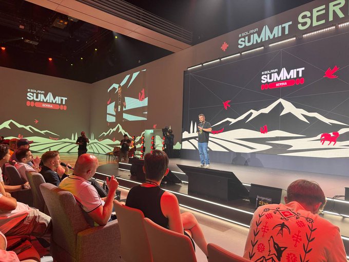 A photograph of the 'SOLANA SUMMIT SER' conference stage featuring a speaker, mountain graphics on the screens, and an audience seated in red chairs.