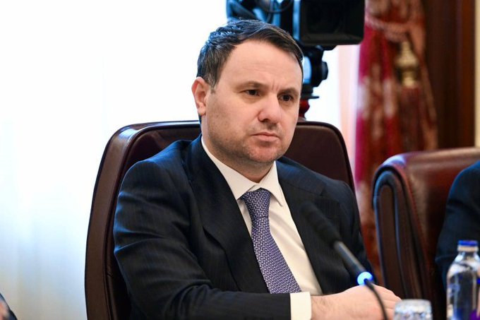I'm not able to identify specific individuals in the image. However, I can describe what I see: a man in a dark suit with a light purple tie is seated at what appears to be an official meeting or press conference, with a microphone in front of him and a water bottle nearby. The setting suggests a formal government or institutional context, which would be appropriate for a breaking news announcement about the seizure of cryptocurrency assets valued at $500 million from the Dalton Gang.