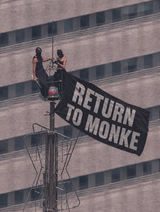 Two protesters dressed in black stand atop a tall structure, unfurling a large black flag with bold white text reading "RETURN TO MONKE." The aerial perspective emphasizes the daring act and makes the large flag's message stand out against the patterned building facade behind them.