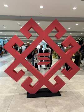 A red geometric sculpture or logo displayed in a lobby or convention center, with people walking in the background.