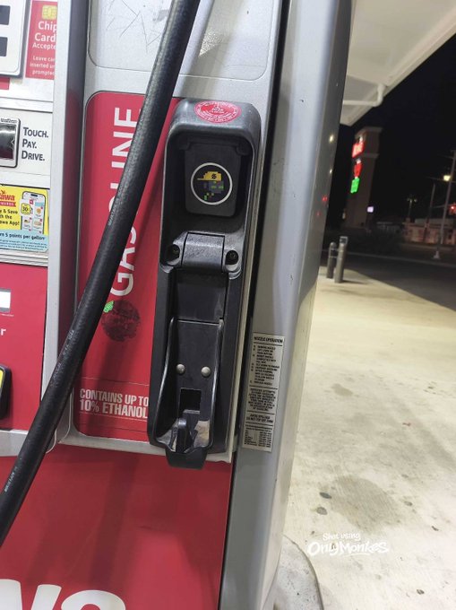 A close-up of a red gas pump nozzle at a gas station at night, labeled with 'CONTAINS UP TO 10% ETHANOL' and 'TOUCH PAX DRIVE'.