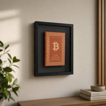 The image features a black frame with an orange book titled "The Bitcoin Standard" displayed inside, which is hanging on a wall next to some books. There are also three other books visible in the scene, one of them being a book about bitcoin. The setting appears to be a living room or a study area where someone might have placed these items for display or reference purposes.