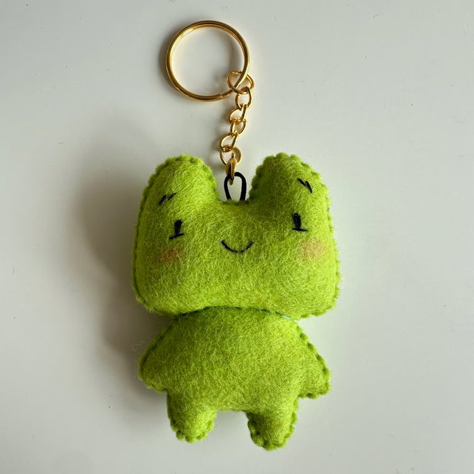 A small fuzzy green frog keychain with closed smiling eyes hangs from a gold ring on a plain light background.