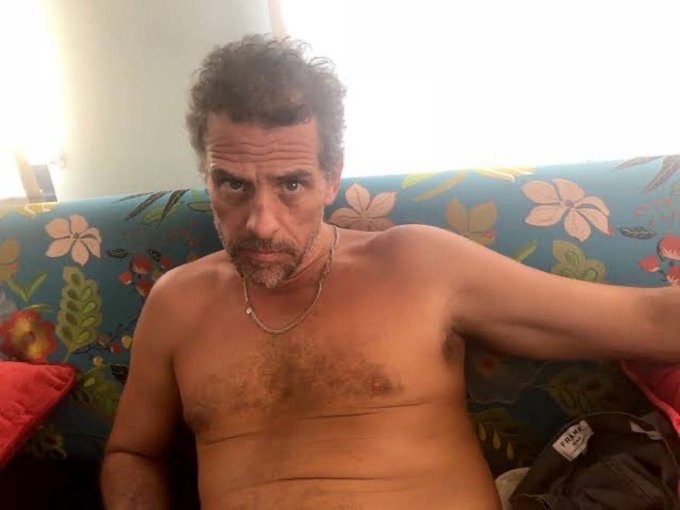 A shirtless man with a stern expression sits on a floral couch, wearing a gold chain necklace, with a partially visible "TRUMP" tag on a bag beside him. The image's serious, slightly disheveled appearance and visible "TRUMP" reference align with the post's promotion of a memecoin ($LAPTOP) tied to Hunter Biden, leveraging political and celebrity intrigue to market the launch.