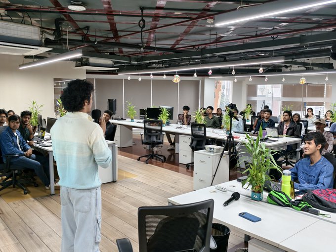 Interior photograph of a modern co-working or training space where a man in a light blue striped shirt stands addressing a group of seated attendees at desks with computers, plants, and equipment.