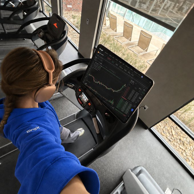 A woman in a blue 'BingX' hoodie runs on a treadmill while watching cryptocurrency charts on a tablet.