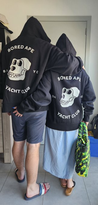 Two people stand with their backs turned, wearing matching black 'Bored Ape Yacht Club' hoodies featuring a skull graphic.