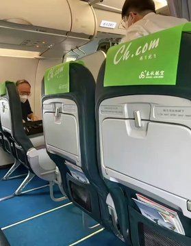 A photo taken from inside an airplane cabin showing rows of seats with green headrests branded 'Ch.com Spring Airlines', with passengers visible.