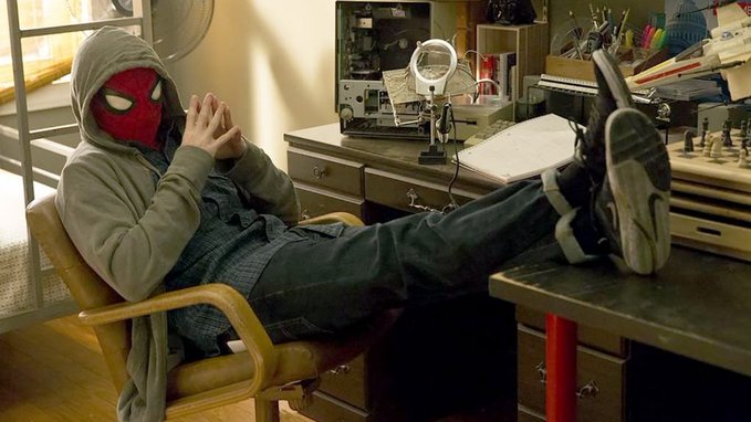 A person wearing a Spider-Man mask and gray hoodie reclines with their feet up on a workbench in what appears to be a workshop or garage, with hands clasped together.