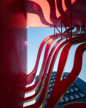 An architectural view looking up at a red metal structure with curved, flame-like slats against a blue sky and city buildings.