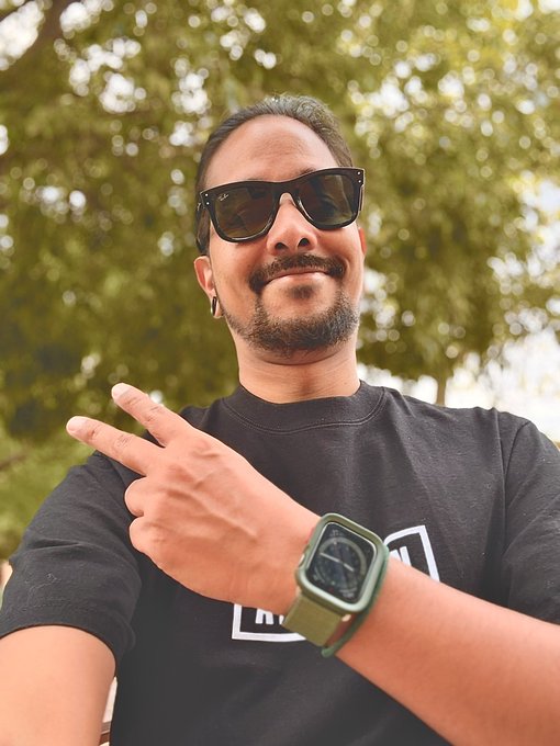 A man wearing sunglasses, a black t-shirt, and a green smartwatch flashes a peace sign outdoors against a leafy backdrop, capturing a cheerful "Good Morning" greeting to his followers.