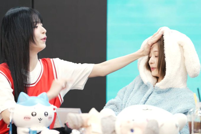 Two women sit at a table; the woman on the left reaches over to adjust the bunny hood of the woman on the right, with several plush toys visible in the foreground.