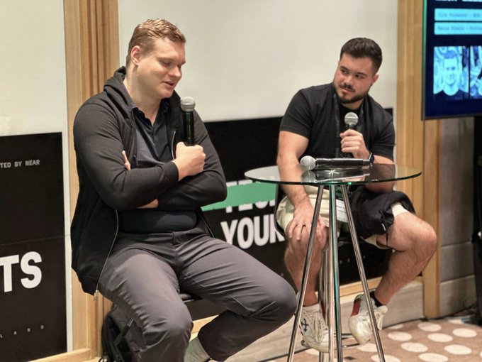 Two men sit on stools holding microphones during a panel discussion, with a partial sign reading 'YOUR' visible behind them.