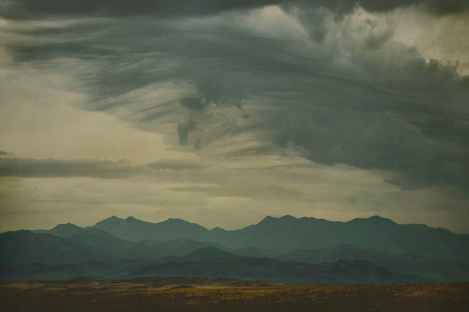 A moody landscape photograph showing layered mountain silhouettes beneath dramatic, sweeping storm clouds with a greenish tint, evoking the atmospheric quality of a painted sky. The image pairs with the post as an artistic visual accompaniment, complementing the caption's "like a painting" sentiment that directs viewers to the linked artwork on objkt.com.