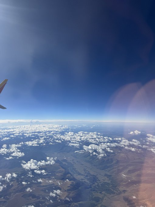 A view from an airplane window showing a vast landscape of mountains, rivers, and scattered clouds below a bright blue sky, with part of the wing visible.