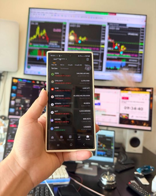 Hand holding a smartphone displaying the vinmart.vn crypto app showing Vietnamese crypto names like PEPE, CHILLOUY, SHIB, BABYDOGE, SERAPH, NOT, SOCIAL, USDC with prices and percentage changes, in front of multiple trading chart monitors.