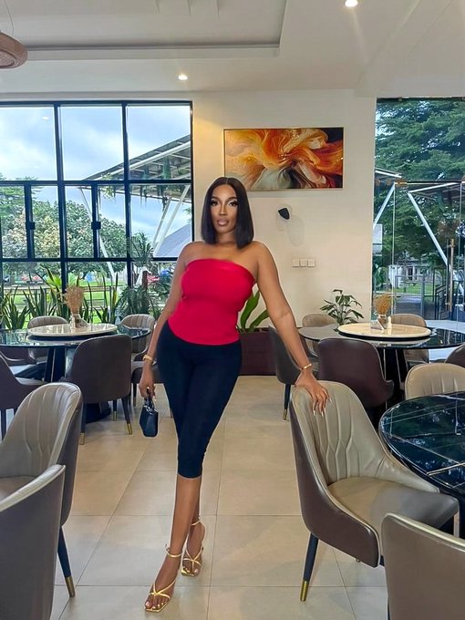A woman in a red strapless top and black pants stands in an elegant restaurant or lounge with large windows, modern furniture, and a floral painting on the wall.