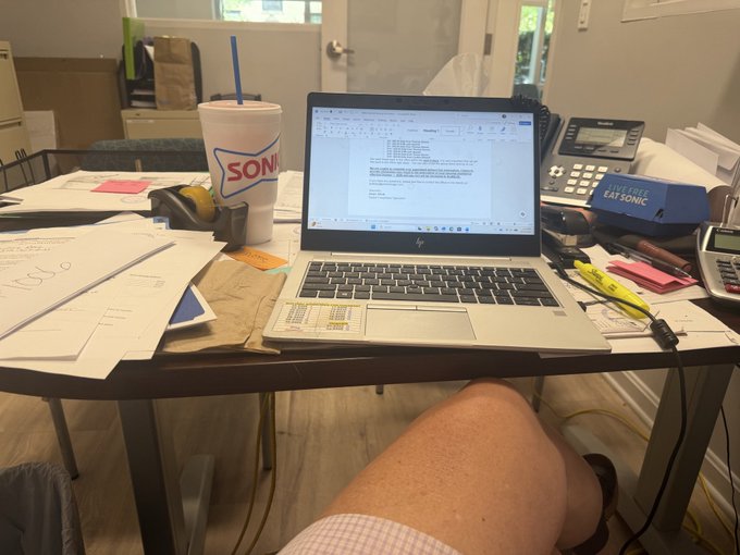 A cluttered work-from-home desk with an HP laptop, a Sonic drink cup, papers, a telephone, and a 'Live Free Eat Sonic' box, with a person's leg in shorts visible beneath the table.