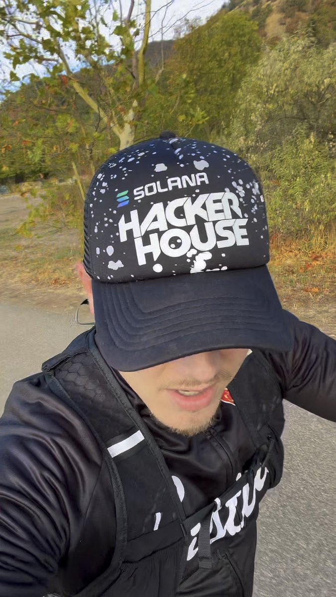 Selfie of a person outdoors wearing a black splatter-pattern trucker hat with 'SOLANA HACKER HOUSE' logo, a black shirt, and a black backpack.