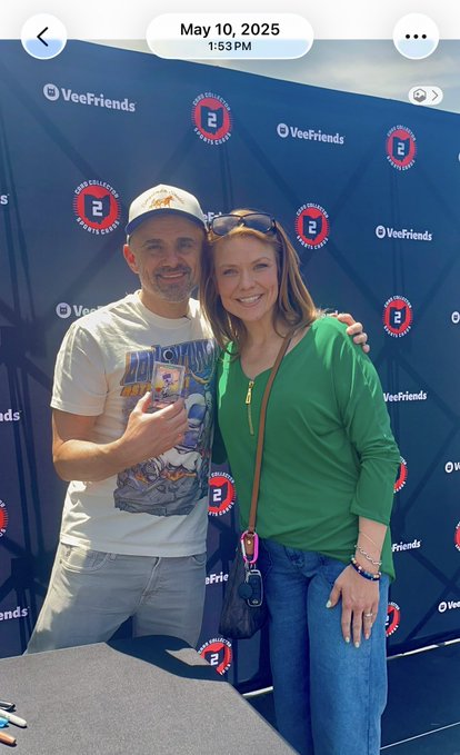 A man and woman posing together at a VeeFriends event on May 10, 2025, with the man displaying a collectible card in front of a branded backdrop.