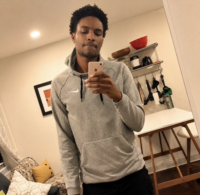 A young man wearing a grey Nike hoodie takes a mirror selfie with a smartphone in a brightly lit room featuring a desk and shelving.