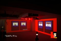 A dimly lit exhibition space with red lighting illuminating three display screens showing images, with Japanese text '「コメディアン」' visible in the corner.