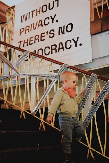 A photograph of a young child climbing a metal staircase beneath a large banner reading 'WITHOUT PRIVACY, THERE'S NO DEMOCRACY.'
