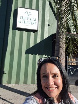 A smiling woman with sunglasses on her head takes a selfie in front of a green striped wall with a sign reading 'THE PALM & THE PINE'.