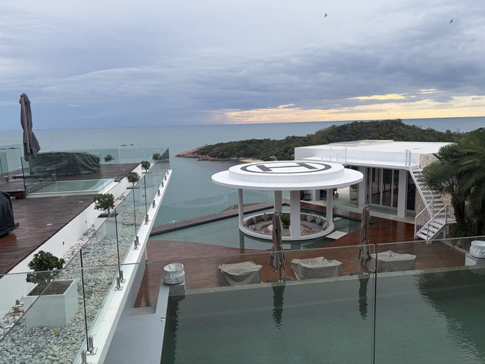 A scenic view of a luxurious coastal property with infinity pools, wooden decking, and a circular helipad structure, overlooking the ocean and a forested headland at sunset.