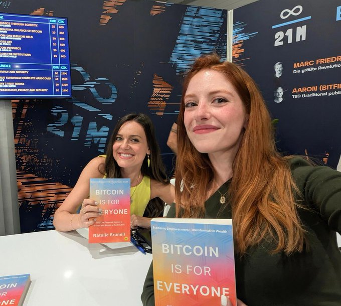 The image features two women sitting at a table, each holding up a book titled "BITCOIN IS FOR EVERYONE!". They are smiling and appear to be promoting or discussing the benefits of Bitcoin as an alternative form of currency. The background includes a TV screen displaying information about Bitcoin, suggesting that they might be at a convention or event related to cryptocurrency.