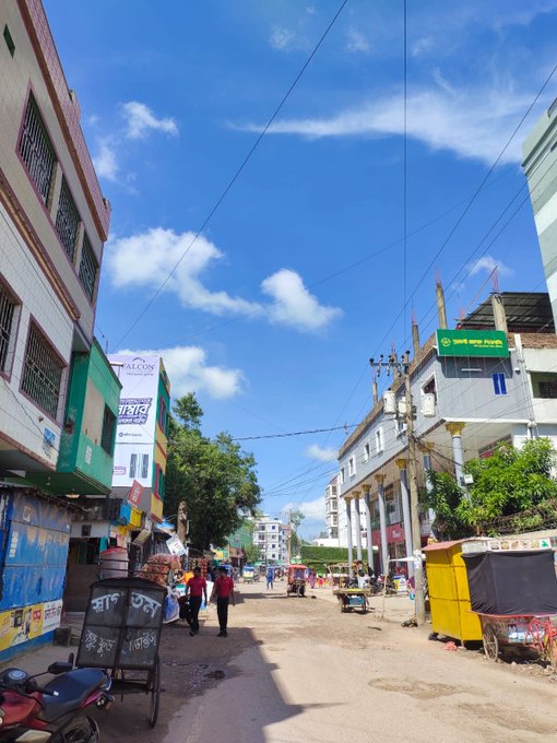 A street view shows a sunny day with blue skies in a town, featuring buildings on either side, power lines, and signs with Bengali script.