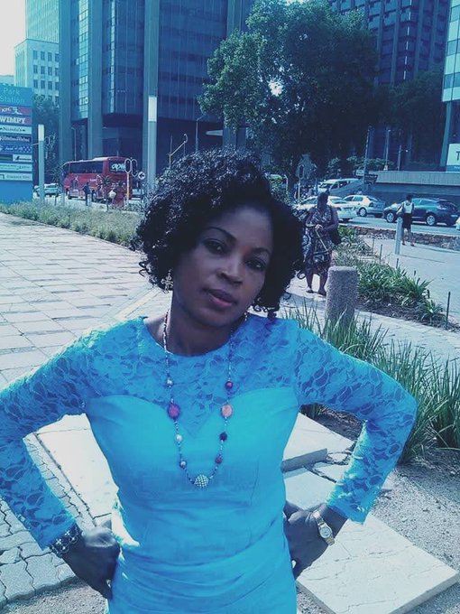 A confident woman poses with hands on hips in front of a modern building, wearing a vibrant blue lace top and colorful beaded necklace, embodying the determined spirit of "grinding harder" and being devoted to a loved one as mentioned in the caption.