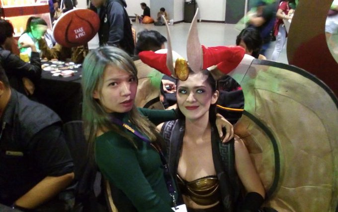Two cosplayers pose together at what appears to be a convention; one is dressed in an elaborate horned demon-style costume with green makeup, while the other wears a green top and a purple lanyard.