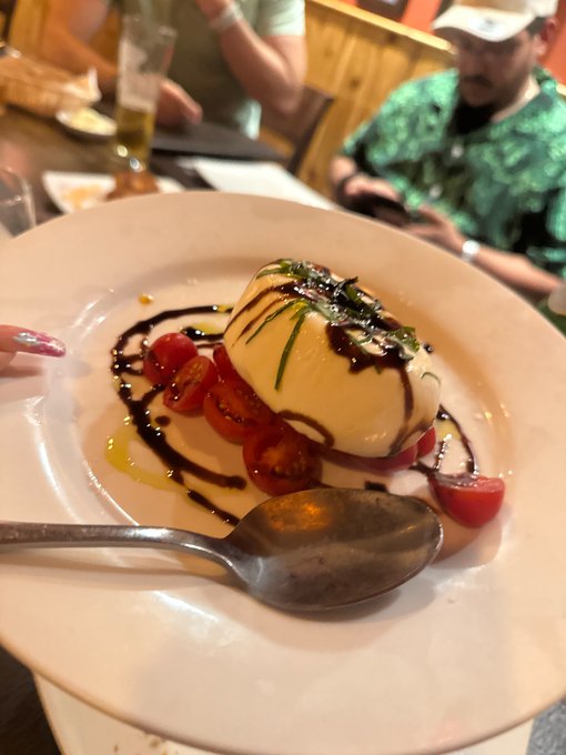 Restaurant photo of a plated burrata or mozzarella dish topped with herbs and drizzled with balsamic glaze, served with cherry tomatoes and a spoon on a white plate.