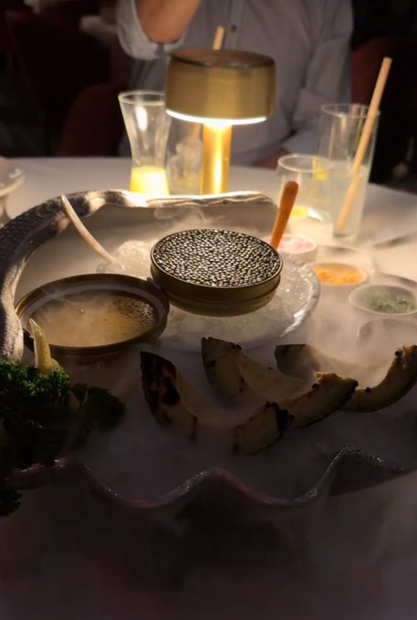 An upscale dining experience featuring an artfully presented seafood course with grilled avocado slices and a decorative shell-shaped bowl, surrounded by molecular gastronomy elements and a small table lamp creating an intimate ambiance. The luxurious, smoky atmosphere complements the post's aspirational tone about pursuing generational wealth and prosperity.
