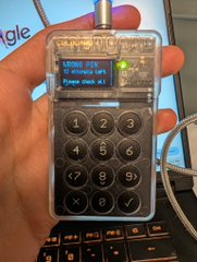 A hand holds a transparent keypad device showing "LOCK_KEYB" and "Please unlock it!" on its small display, with a numeric keypad below. The image humorously relates to the post by showing a hardware PIN entry device, underscoring the need to remember the unlock PINs.