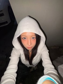 A selfie of a young woman with long brown hair wearing a white hoodie pulled up over her head, posing indoors.