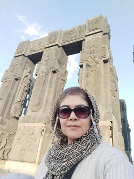 A selfie of a woman wearing sunglasses and a black-and-white patterned headscarf, standing in front of ancient Egyptian ruins with carved hieroglyphs under a blue sky.