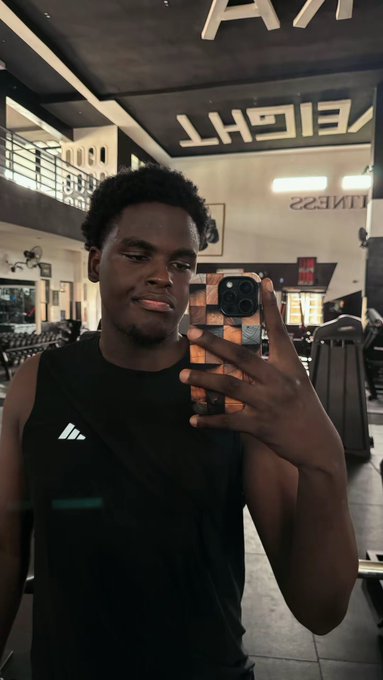 A young man in a black Adidas tank top takes a mirror selfie in a gym, with "WEIGHT" and "FITNESS" text visible in the mirrored reflection, capturing a casual "Late gm" greeting post-gym session.