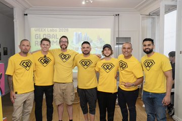 The image shows a group of six men standing together with their arms around each other's shoulders, all wearing yellow t-shirts that say "weex lab". They are posing for a photo at an event or gathering. The background features a projector screen displaying the text "Global Labels 2028" and "WEEX Lab".