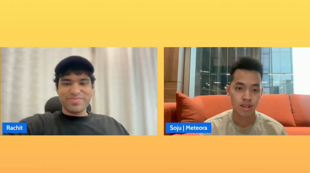 The image shows two men sitting next to each other, both wearing black shirts, and they are talking into microphones. The background of the image features an orange couch placed against a yellow wall.