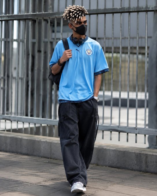 A young man with dreadlocks walks down a city street wearing a light blue Manchester City Puma soccer jersey, black mask, headphones around his neck, and black cargo pants.