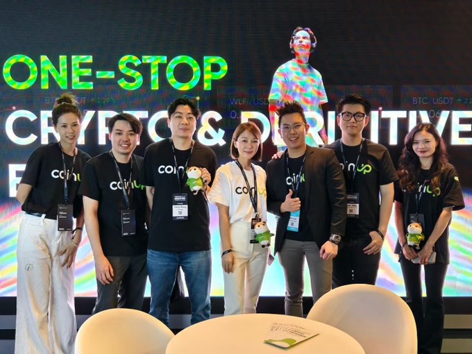 A team wearing "coinup" branded shirts poses together at an event with a large backdrop reading "ONE-STOP CRYPTO & DERIVATIVE," visually aligning with the message of collaborative pride in building together.