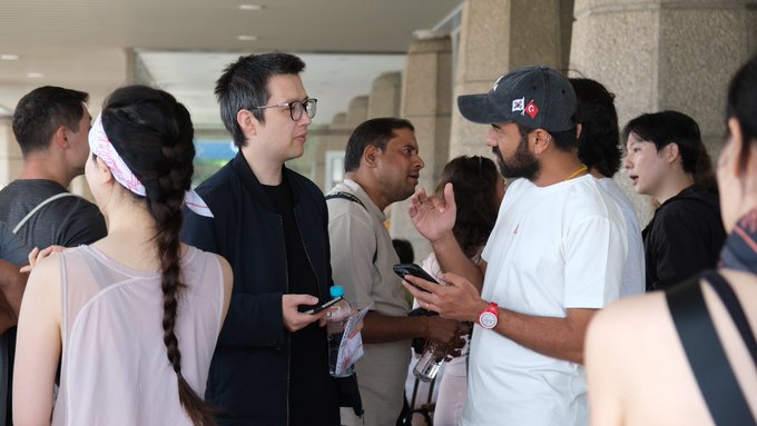 A diverse group of people are gathered together indoors, with two men at the center appearing to be in animated conversation—one wearing a navy jacket and glasses, the other in a black cap and white t-shirt gesturing with his hand while holding a phone. The scene captures a casual networking moment, fitting the post's context of an in-person gathering of blockchain/crypto community members (Avax team and Tiger Research) on their first day in Korea.