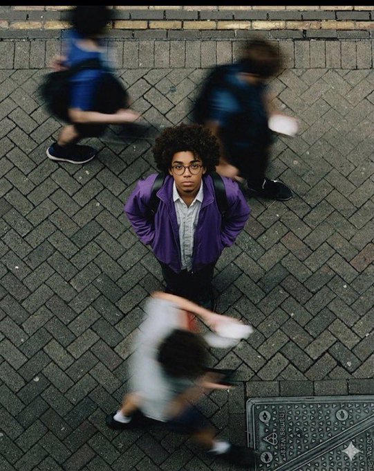 A top-down photograph of a person with an afro, glasses, and a purple jacket standing still on a paved street while blurred figures rush past them, creating a stark contrast between stillness and motion.
