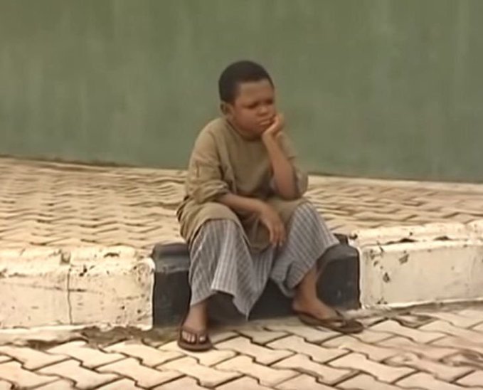 A young boy sits on a suitcase on a paved surface, resting his chin on his hand with a contemplative expression.