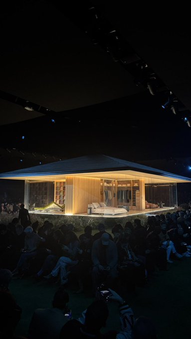 A nighttime architectural rendering of a modern, flat-roofed pavilion with warm interior lighting, displayed to an audience in a dark auditorium, showcasing a bedroom and what appears to be a hammock.