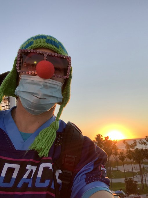 A person wearing a green knit cap with earflaps, a light blue surgical mask, and pixelated sunglasses with a red clown nose takes a selfie at sunrise. They wear a purple jersey with partial text reading "DACS" visible, aligning with the post's reference to "GM" and "CryptoPunks" with a quirky, clown-like aesthetic matching their playful "DEGEN" vibe.