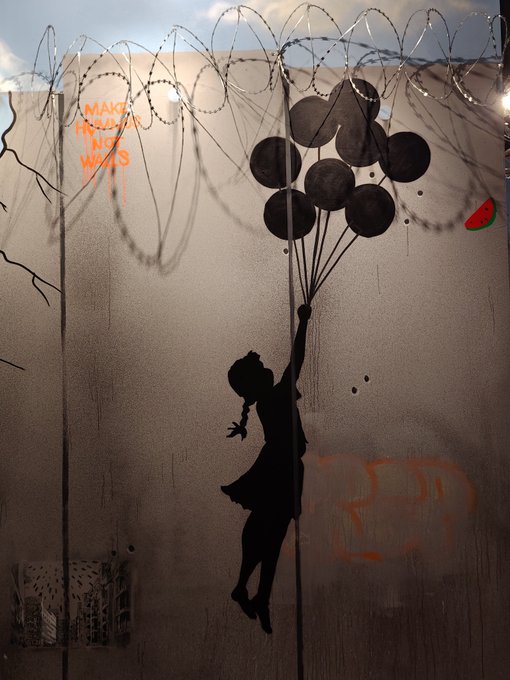 A Banksy-style street art mural depicting a girl silhouette floating with balloons toward razor wire on a wall, with the text 'MADE FOR THE WALLS' and a small watermelon graphic.