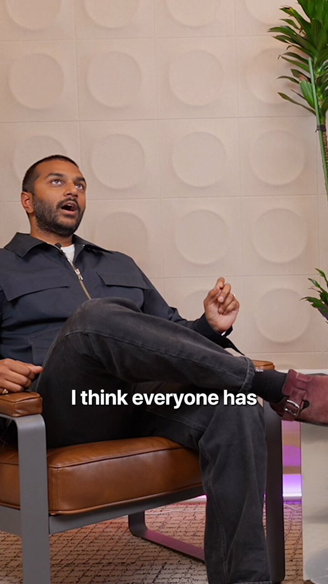 The image shows a man sitting in an office chair with his legs crossed, wearing a blue jacket and black pants. The background features a white wall with circular shapes, giving the impression of a modern workspace or office environment. There are also two potted plants present in the room - one on the left side and another on the right side.